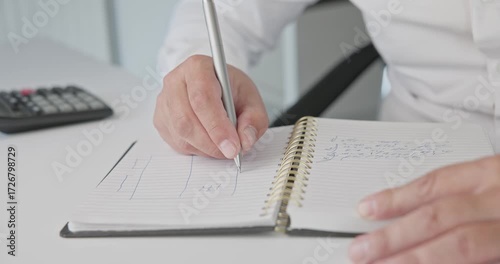 Close-up of hands - a man in a shirt draws a diagram and takes notes with a pen in a notepad on a metal spring on an office desk