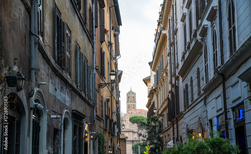 Canvas Print Narrow alley in central Rome with tall historic buildings, shuttered windows, and classic iron lanterns, leading to a distant church tower surrounded by terracotta rooftops and stone pine trees