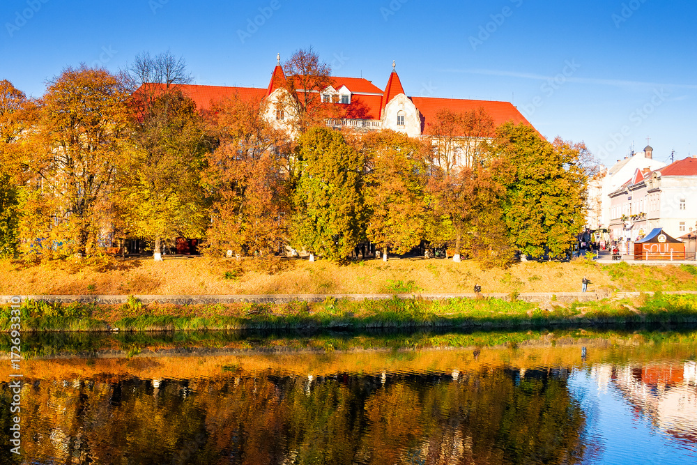 Naklejka premium uzhhorod, ukraine - 31 oct, 2010: embankment of the river Uzh in autumn. beautiful view of old city with colorful foliage on trees in evening light. scenic european downtown in fall season