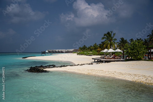 Fototapeta Naklejka Na Ścianę i Meble -  White sandy beach on a Maldivian island with turquoise water, over water villas in the distance, palm trees, sun loungers with umbrellas, and volcanic rocks under a dramatic tropical sky.