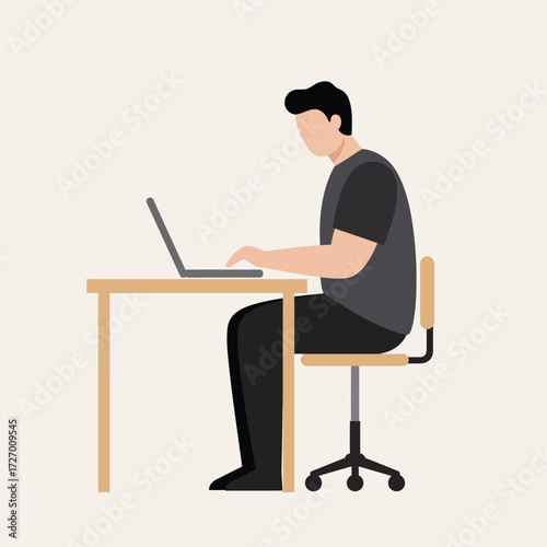 A man in a dark t-shirt and black pants sits at a wooden desk, typing on a silver laptop, with a swivel chair.