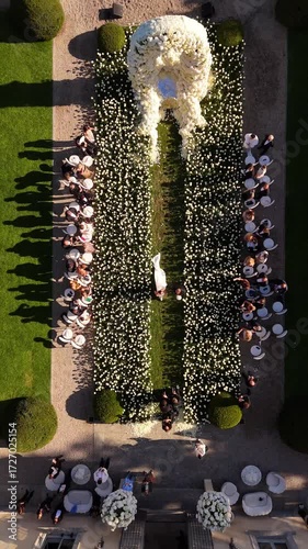 Cinematic drone view of bride walking with her father to floral arch altar, guests on both sides, fresh roses along aisle, Lake Como, Italy. Elegant lakeside wedding ceremony, luxury event