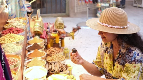 Tourist woman in floral shirt and hat exploring spice market stall in Dubai heritage bazaar, she is smelling and selecting colorful spices and herbs, teas on display 