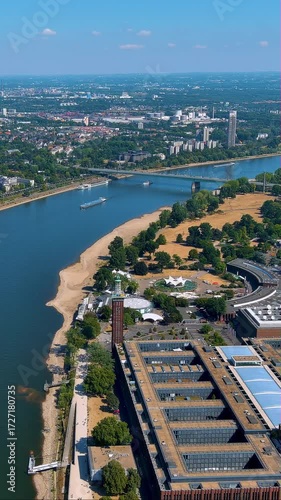 4K vertical drone video sweeps Cologne’s Rhine waterfront: Koelnmesse halls, Deutz beach park, bridge and passing ships under clear summer sky - vibrant footage for travel and trade - fair stories.