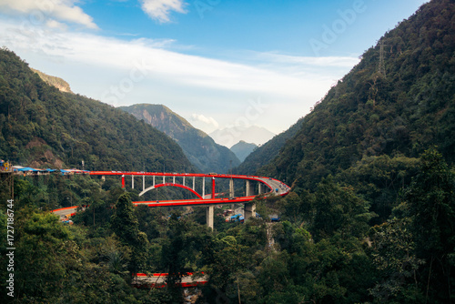 A vibrant red bridge snakes through lush green mountains under a clear blue sky.  Kelok 9, or the 9 hairpins, Harau, West Sumatra, Indonesia
