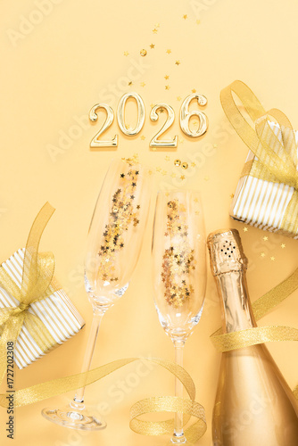 Happy New Year 2026! Bottle of golden champagne and glasses, elegant gifts with ribbon and bow and golden confetti on golden background.