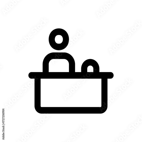 A simple black and white icon depicting a person standing behind a desk with a circular object on it