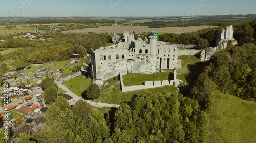 Aerial drone footage circling Ogrodzieniec Castle in Poland, highlighting the medieval fortress, stone walls, towers, and surrounding scenic landscapes from all angles.