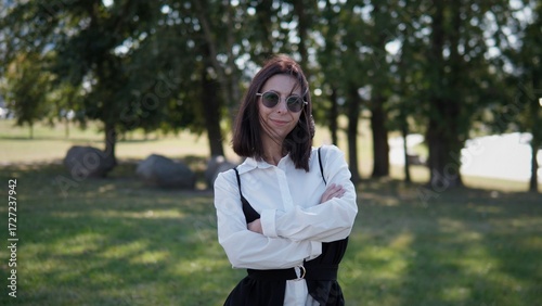 A business woman stands confidently outdoors with arms crossed, showcasing a stylish outfit. The lush greenery in the background adds a serene touch to her strong presence