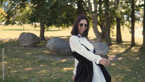 A confident businesswoman stands in a park, showcasing her stylish outfit. Surrounded by trees and large rocks, she smiles as she poses playfully for the camera under the warm sunlight