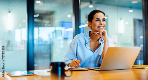 Smiling woman in office working on laptop and looking out with confidence