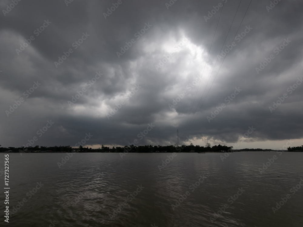 Fototapeta premium Dramatic dark storm clouds gathering over a tranquil river before a thunderstorm, creating an ominous and moody atmosphere