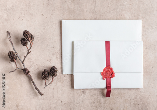 White envelope sealed with red ribbon and boho decoration on beige stone background and dry plant. Stationery packed in a box and ready to mail