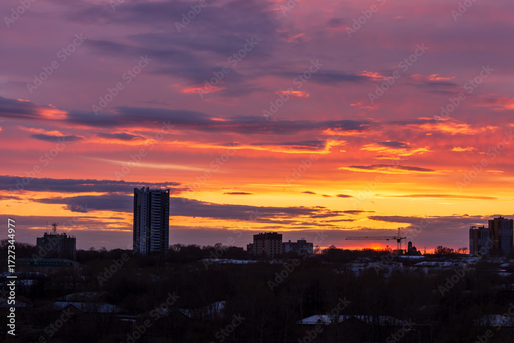 Naklejka premium A vibrant sunset over a city skyline. The sky is filled with shades of orange, pink, and purple. Silhouettes of buildings are visible against the colorful backdrop.