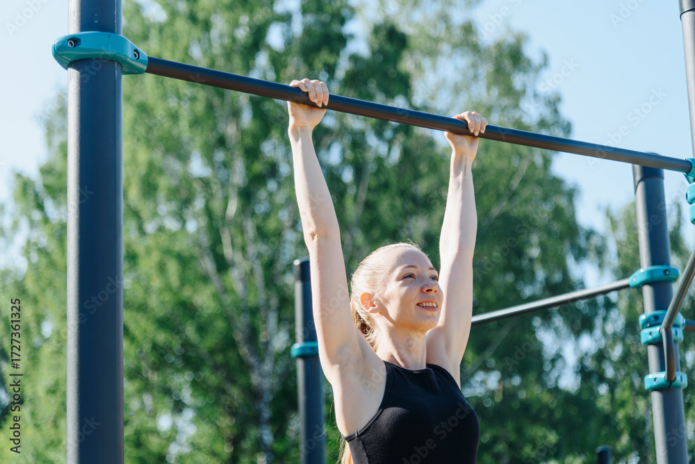 Fototapeta premium young sportive caucasian woman doing exercises on horizontal bar outdoors in summer, beautiful redhead girl, black sportswear, healthy lifestyle concept