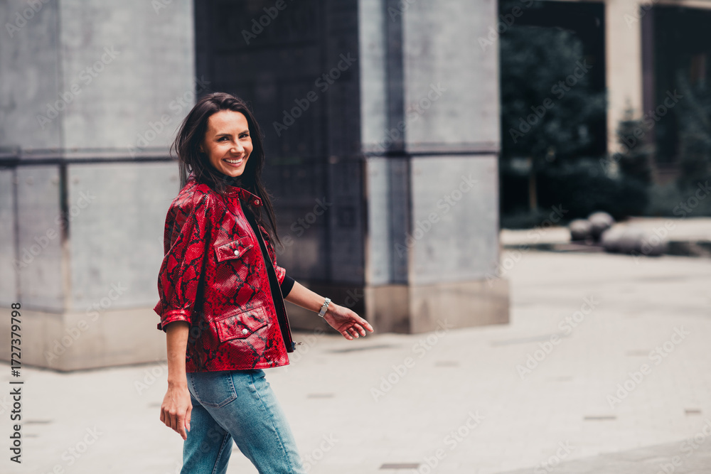 Fototapeta premium Beautiful young woman in a red jacket walking outdoors in a city during daylight, showcasing casual urban fashion