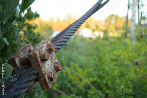Close-up of a rusty steel cable clamp holding a tensioned metal rope against a blurred green natural background. Concept of strength, durability, industrial detail, and aging metal outdoors.
