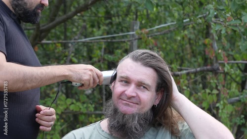 A bearded man with a pigtail cheerfully nods his head while being shaved with a hair clipper outdoors, expressing humor and positive emotions.