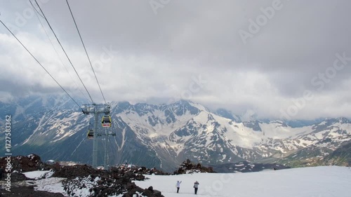 Scenic view of ski lift ascending through snowy mountains under cloudy sky. Two people are seen standing on the snow-covered slope, surrounded by rugged peaks and dramatic alpine scenery