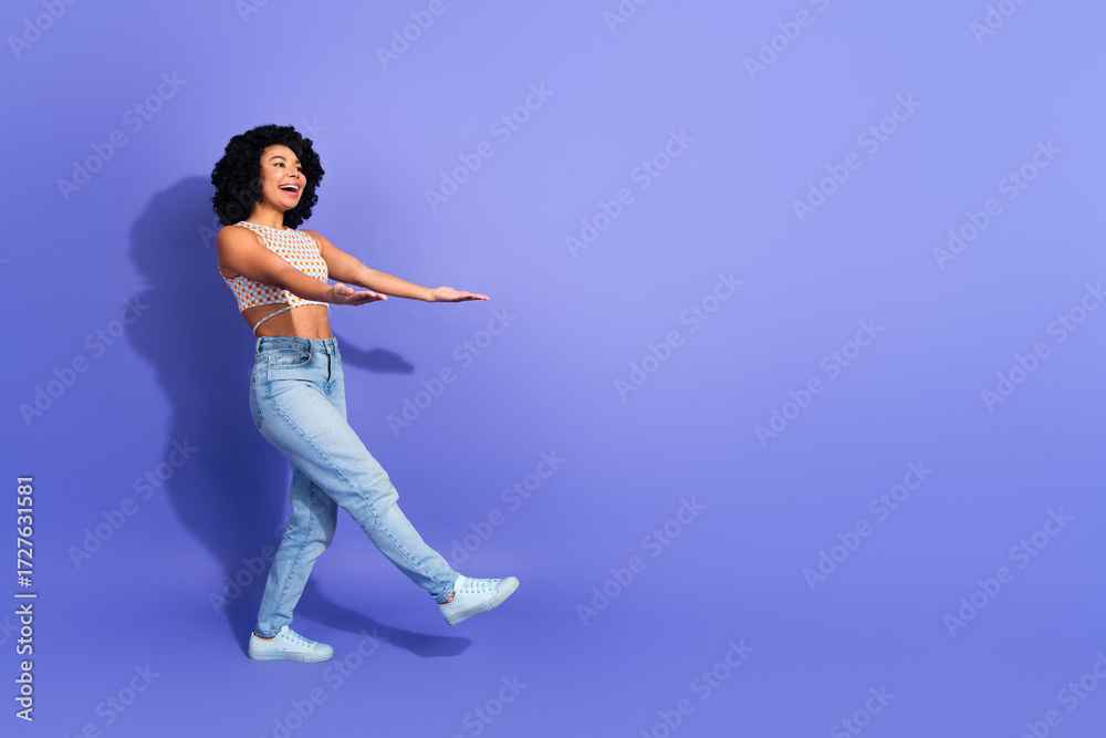Obraz premium Cheerful young woman with curly hair dancing in a colorful casual outfit against a vibrant purple background
