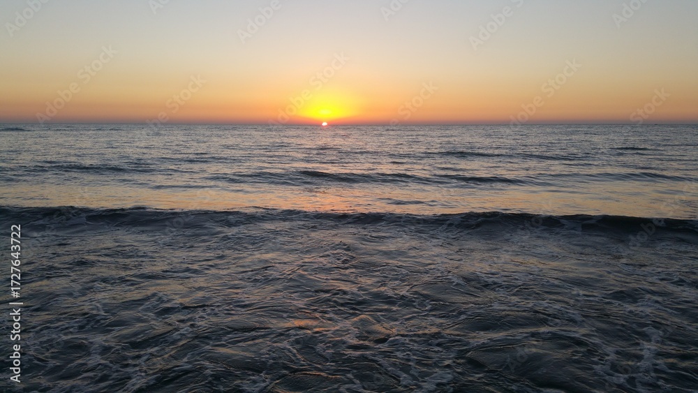 Obraz premium Sunset over the ocean horizon casting golden light across the water on a calm evening at the beach