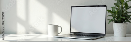 Modern laptop with blank screen sits on white desk next to coffee mug and green plant. Bright window light casts shadows on wall. Minimalist workspace setup for remote work.