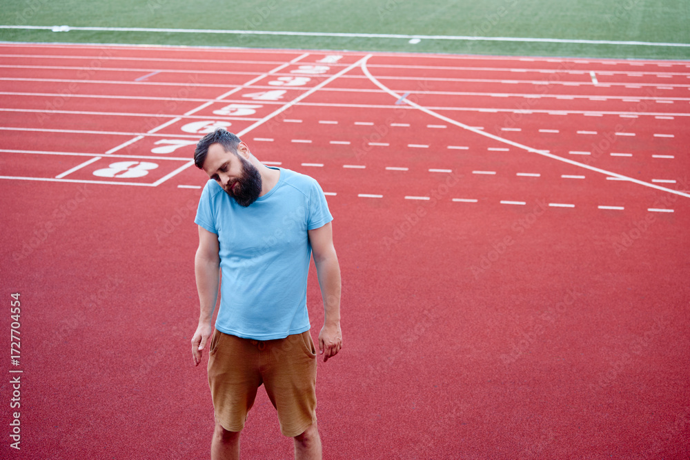 Obraz premium A casual man in a blue shirt and shorts stretches neck on an outdoor running track on a sunny day. The red track and green field provide a contrasting and vibrant background.