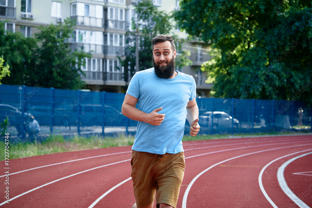 Obraz premium A bearded man wearing a light blue shirt jogging on a red track with urban buildings and lush greenery in the background, enjoying a healthy and active lifestyle outdoors.