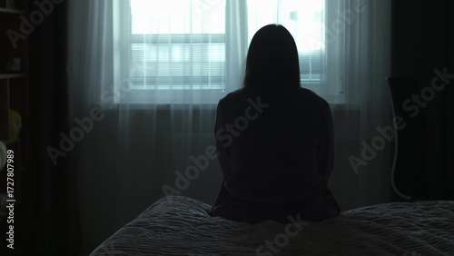 Back view Silhouette of a millennial woman sitting on a bed in the dark bedroom, depression, pain. Portrait of Unhappy sad woman sitting against window and thinking about her life