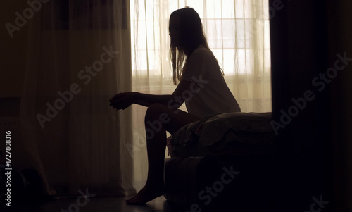 A woman sits alone on the edge of a bed in an empty room, her silhouette framed by soft light. She appears lost in thought, emotionally overwhelmed, facing a personal mental health crisis.
