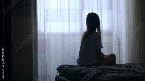 Silhouette of unrecognized woman sitting on a bed in the dark bedroom, depression, pain. Portrait of Unhappy sad woman sitting against window and thinking about her life