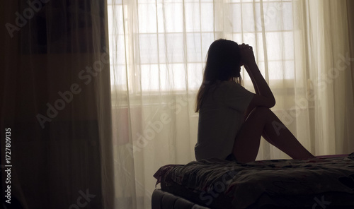 A woman sits alone on the edge of a bed in an empty room, her silhouette framed by soft light. She appears lost in thought, emotionally overwhelmed, facing a personal mental health crisis.