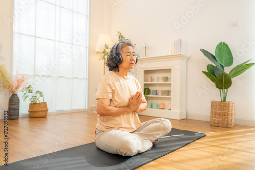 Happy Asian senior woman meditating in lotus pose with sound therapy on headphones at home. Healthy elderly woman enjoy wellness healthcare lifestyle relaxing, meditation yoga exercise in living room.