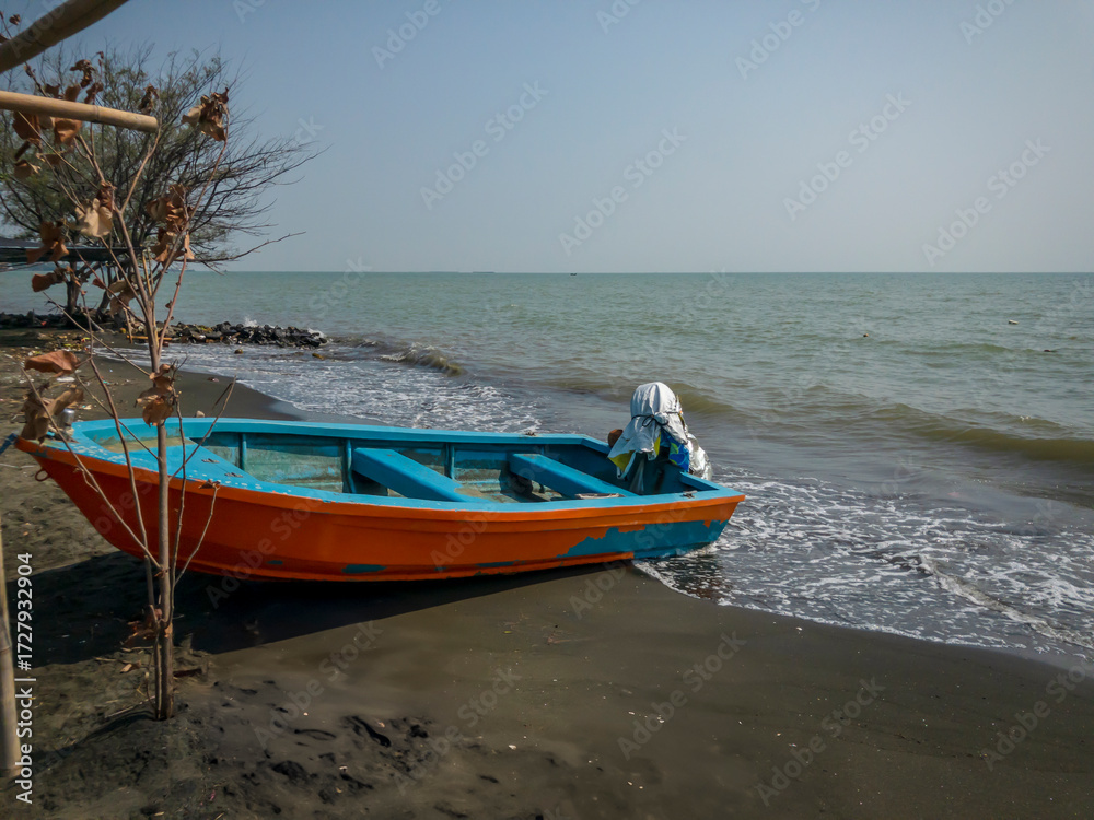 Fototapeta premium A colorful orange and blue wooden fishing boat rests on the sandy shore as gentle waves touch the coast under a clear sky