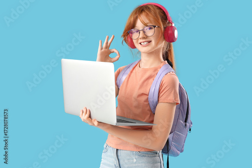 Cute schoolgirl with modern laptop, backpack and headphones showing OK on blu...