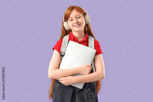 Cute schoolgirl with modern laptop and headphones on lilac background