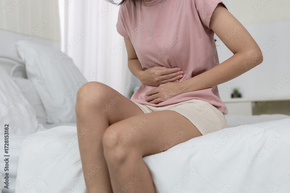 Obraz premium women touching her lower abdomen with discomfort while sitting on bed, depicting menstrual cramps, stomachache, digestive issues, or abdominal pain in home health or lifestyle concept.