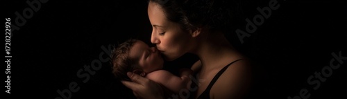A loving mother gently kisses her sleeping newborn baby on the forehead in a tender moment against a dark and moody background.
