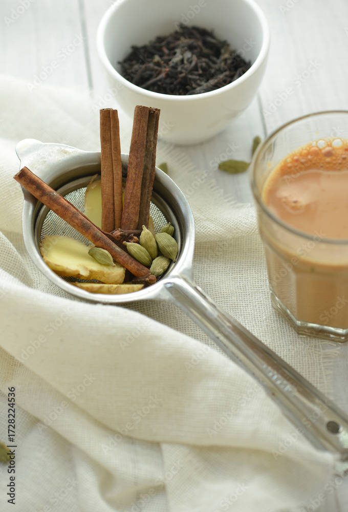 Fototapeta premium Ingredients for Indian Masala Chai or Tea. Fresh ginger, cinnamon and cardamom are placed in a tea sieve along with a hot cup of Chai.