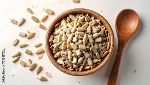 Overhead view of peeled sunflower seeds piled high in a small wooden bowl next to a wooden spoon on a white background.