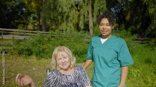 Portrait of positive attractive elderly woman in wheelchair and African American female caregiver in uniform friendly talking and sharing, looking at scenic view during morning walk in summer park.