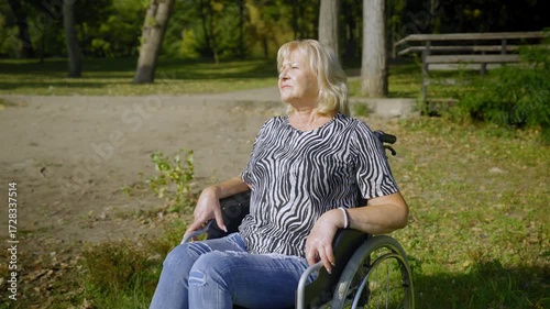 Joyful attractive senior female wheelchair user spending active lifestyles , inhaling fresh air and relaxing in summer nature , expressing happiness and positivity .
