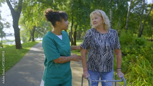 Positive beautiful elderly woman on retirement with mobility walker and caring pretty African American female carer in uniform talking , enjoying leisure and fresh air in public park.