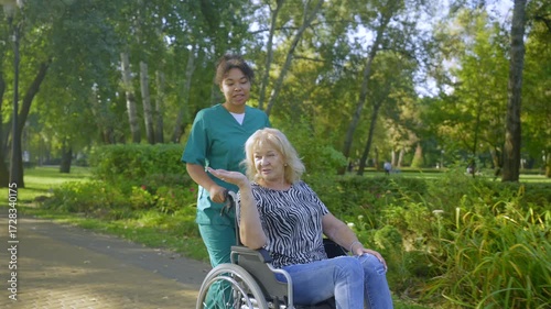 Positive charming African American female caregiver in uniform friendly talking with happy elderly woman with age-related health conditions, rolling wheelchair in summer park.