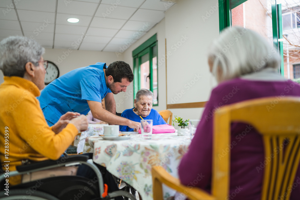 Obraz premium Caregiver helping an elderly woman in a blue shirt with an activity at a table in a bright common area of a nursing home