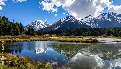 A tranquil scene, mountains reflect in a still lake under a blue sky with fluffy white clouds. The foreground holds marsh grasses