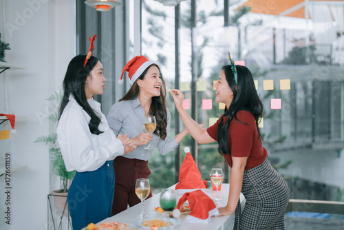 Three Asian businesswomen laughing and toasting at a festive office Christmas party, wearing hats, enjoying snacks, drinks and warm holiday camaraderie in a modern workplace