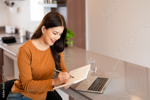Young woman  sitting at counter bar in kitchen at home or apartment and makes notes in notebook writing down general ideas, schedule and planning .