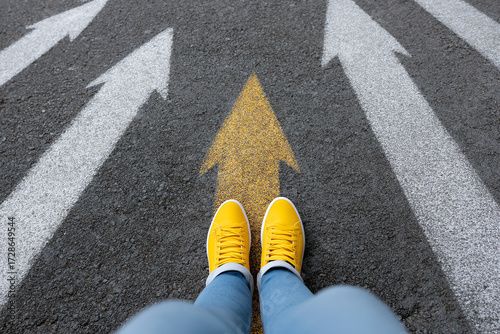 Person wearing yellow shoes stands on a road with multiple white arrows and one yellow arrow pointing forward, symbolizing direction, choice, and moving ahead