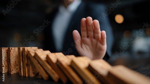 A person in business attire stops a falling row of wooden dominoes with their hand, symbolizing intervention, prevention, or control in a chain reaction